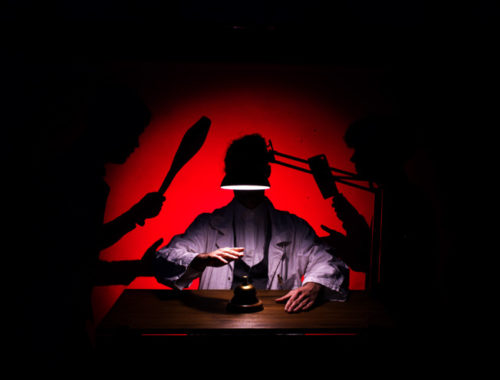 Scène de spectacle avec un personnage éclairé sous une lampe, dans une ambiance rouge et noire évoquant une quête de paix intérieure.